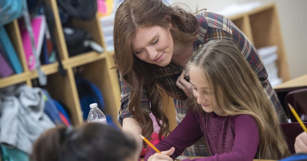 Student teachers helps elementary student at her desk.