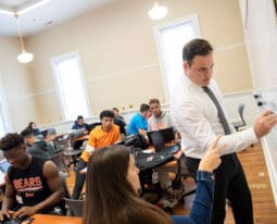 Professor writes on whiteboard in front of business class.