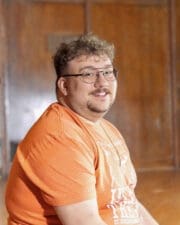 Person with short curly hair, glasses, and an orange T-shirt smiling while seated indoors against a wood-paneled background.