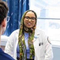 Student Doctor providing healthcare to a patient at the Bear Health Clinic. The student doctor is wearing a green, hijab-style headscarf in a light blue room with dark blue curtains
