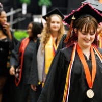 Graduates walking after commencement. The woman in the foreground has a medal and chords around her neck. All the graduates are wearing a black cap and gown.