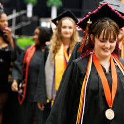 Graduates walking after commencement. The woman in the foreground has a medal and chords around her neck. All the graduates are wearing a black cap and gown.