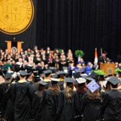 Undergraduate Commencement. Black backdropped stage with a crowd of students in black cap and gowns, looking at the stage with the deans present.