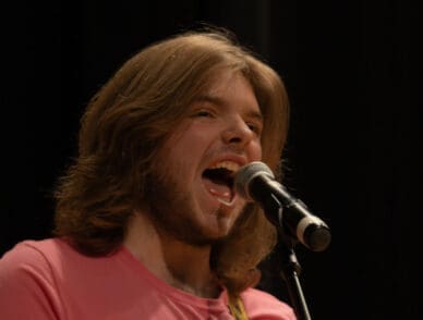 "A young man with long, light brown hair passionately sings into a microphone on stage. He wears a bright pink shirt and has an expressive, open-mouthed pose, emphasizing his vocal performance. The background is dark, drawing focus to the performer.