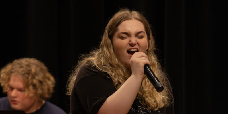 A student passionately sings into a microphone during a live performance. She has long, curly blonde hair and wears a black shirt. A pianist is visible slightly behind her on the left.