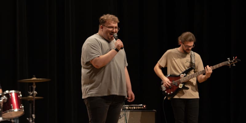 A man in a gray t-shirt stands on stage holding a microphone and smiling while addressing an audience. Beside him, another man in a matching gray shirt plays an electric bass guitar. A red drum set is partially visible on the left, and a black curtain provides a backdrop for the performance setting.