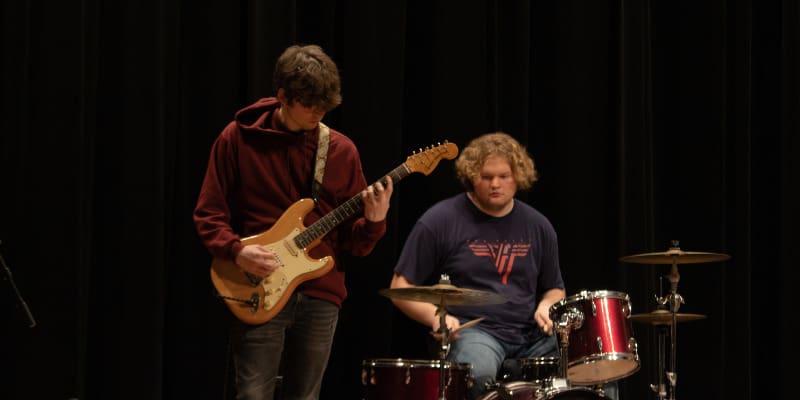 Two students perform on stage against a black curtain backdrop. One student in a maroon hoodie plays an electric guitar, while the other, wearing a dark blue Van Halen t-shirt, plays a red drum set. Both musicians are focused on their instruments during the live performance.