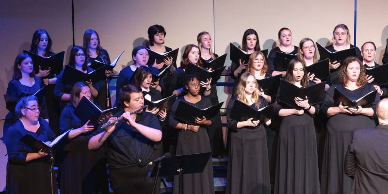 A choir of students dressed in black sings on risers during a performance. They hold black folders and are arranged in rows, with a flutist performing in the front left of the group.