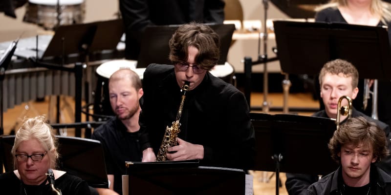 A concert band performs on stage. A young man in the center plays a saxophone solo, standing among other seated musicians. Everyone is dressed in black, and percussion instruments are visible in the background.