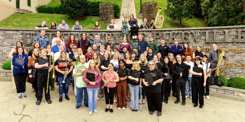 A large group of UPIKE students and faculty pose for a group photo in front of a stone wall engraved with "University of Pikeville." Some hold musical instruments, while others wear matching black performance shirts. The setting is a landscaped outdoor area with greenery and stairs behind the group.