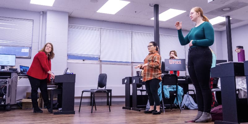 A classroom scene shows several students and an instructor engaged in a music theory activity. Students stand next to desks, gesturing with their hands, while the instructor observes. The room has overhead lights, large windows, and computer workstations.