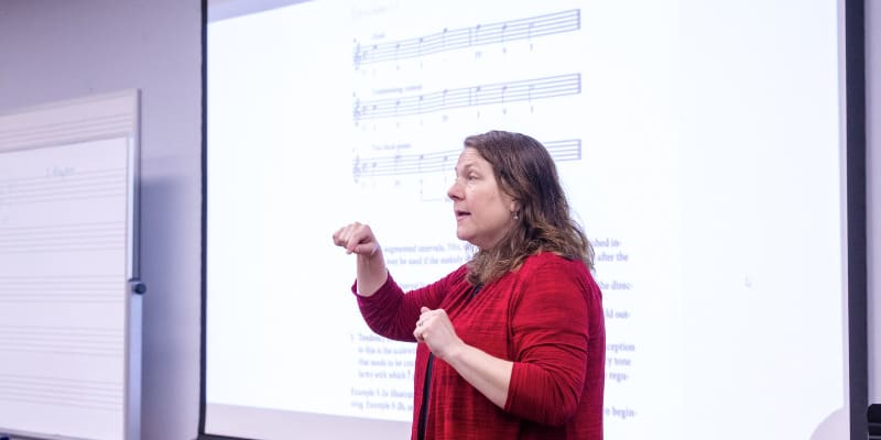 A woman in a red top is standing in front of a large projection screen, using hand gestures as she teaches a music theory lesson. The screen displays musical notation and text, while a whiteboard with staff lines is visible on the side wall. She appears engaged and expressive, actively explaining the material to a class.