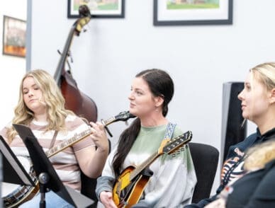 Three students sit in a music classroom holding string instruments, including a banjo and a mandolin. One student is mid-conversation while the others listen attentively. A double bass rests against the wall in the background.