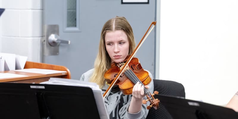 A student with long blonde hair is seated, focused on playing a violin in a rehearsal room. She wears a gray sweatshirt and reads sheet music placed on a black music stand in front of her.