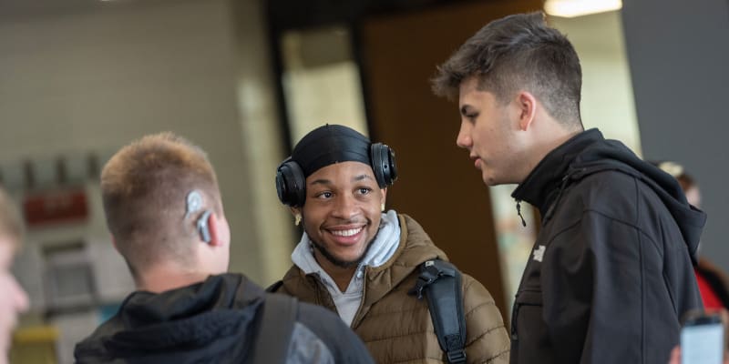 Three male students stand in conversation in a campus hallway. One student smiles brightly while wearing a brown jacket, over-ear headphones, and a black headwrap. Another student with a visible hearing aid is seen from behind, while the third faces the group with a dark jacket. The atmosphere is casual and friendly.