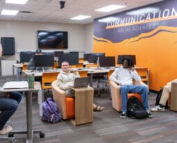 Students sit in a classroom with orange and gray decor under a wall sign reading 'Communication Social Sciences.' Some students use laptops while others engage in discussion. The room features modern seating, individual desks, and computer stations in the background.