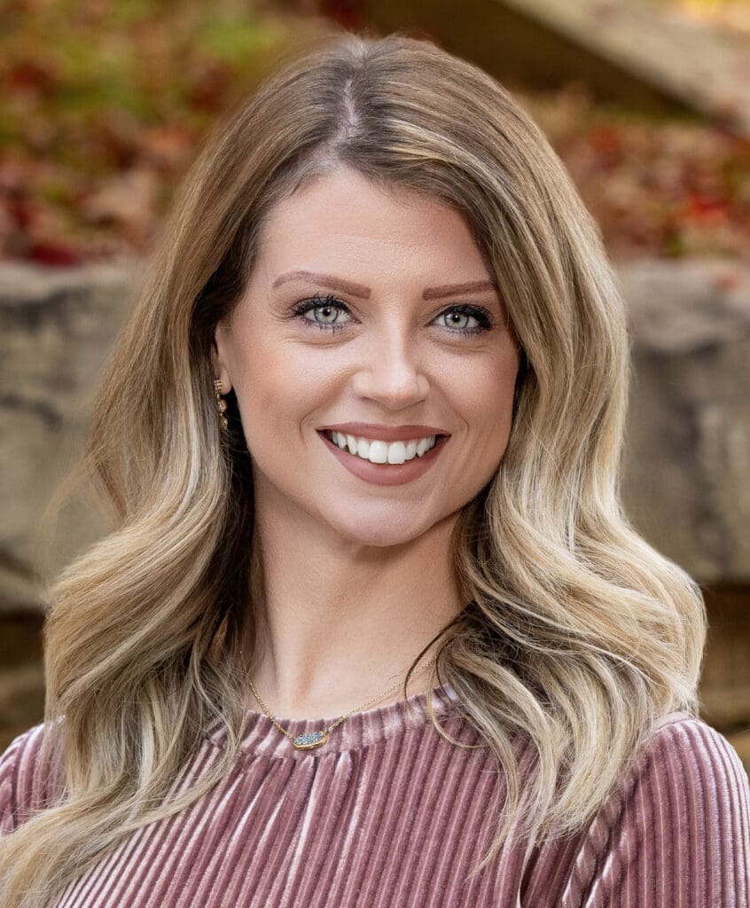 Professional headshot of Jami Slone. She is smiling and facing the camera, with long, wavy blonde hair parted to the side. She is wearing a pink velvet-textured top and a delicate gold necklace with a small, decorative pendant. The background is softly blurred with autumn leaves and stone elements.