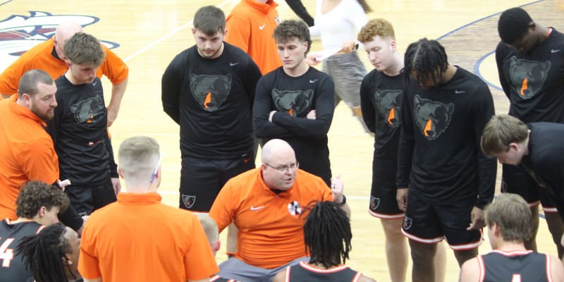 Basketball coach in an orange shirt kneeling and speaking to players in black uniforms with bear logos, gathered in a huddle on the court.
