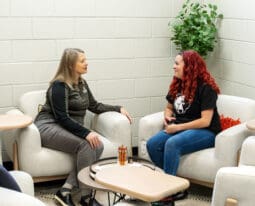 Two women sit across from each other in a lounge area, smiling and talking. One wears a UPIKE bear logo T-shirt, and the other is in a dark top. A small table with pens and notepads sits between them.