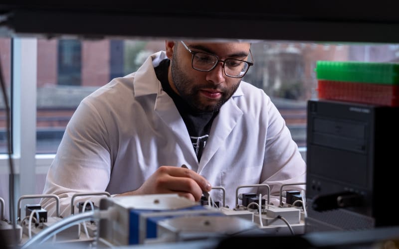 A person in a white lab coat focuses on a small scientific device while working at a lab bench.