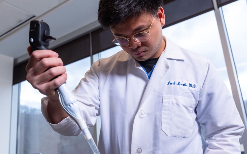 A person in a white lab coat uses a pipette in a science lab with large windows in the background.