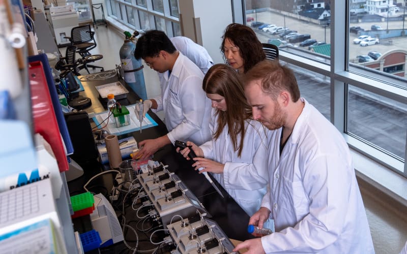 A group of four researchers in lab coats work together at a long laboratory bench with various instruments and containers near tall windows.