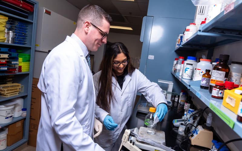 Two people in white lab coats work together at a lab bench with bottles and equipment, both smiling while performing an experiment.